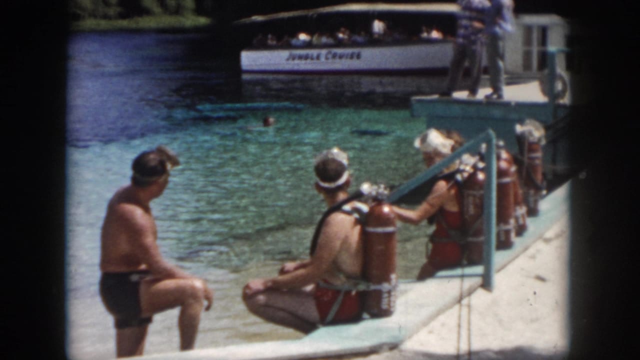 1958 home-movie frame from New Orleans showing visitors in swimwear with red scuba tanks on a concrete dock beside clear water.