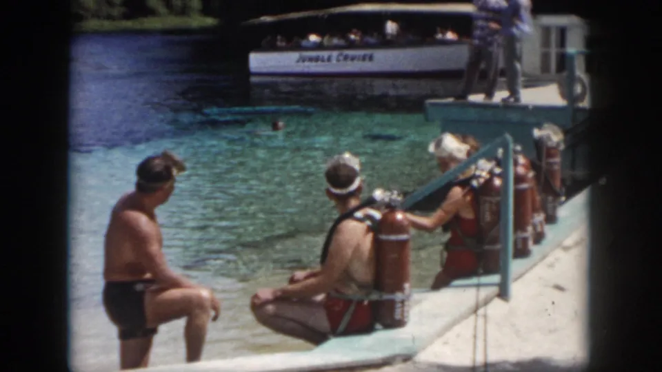 1958 home-movie frame from New Orleans showing visitors in swimwear with red scuba tanks on a concrete dock beside clear water.