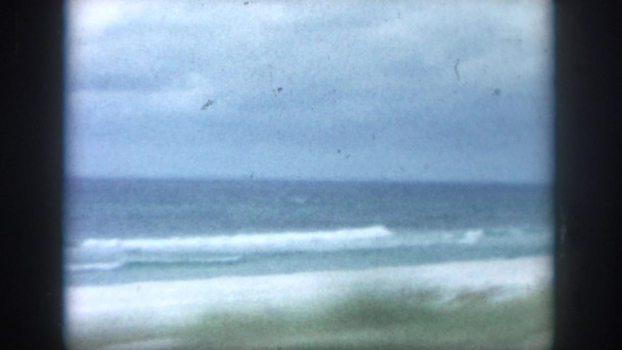 Soft ocean view with pale sand, low surf, and a dark horizon under a cloudy sky.