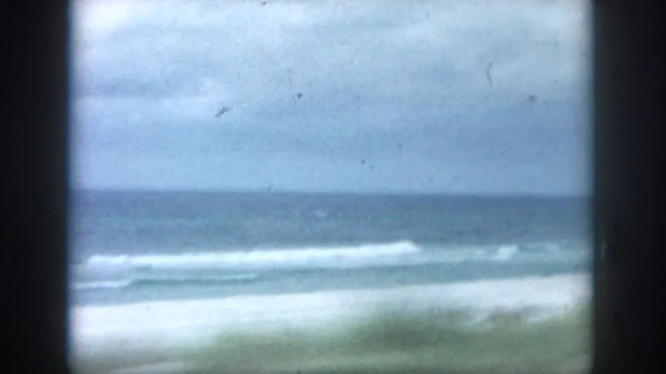 1958 8mm coastal view from the New Orleans, Louisiana metadata set, with soft surf rolling toward pale sand beneath an overcast sky.