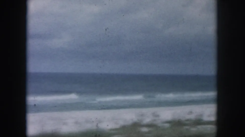 1958 8mm home-movie frame showing a muted beach, distant ocean, soft wave line, and overcast sky, consistent with the coastal travel metadata.