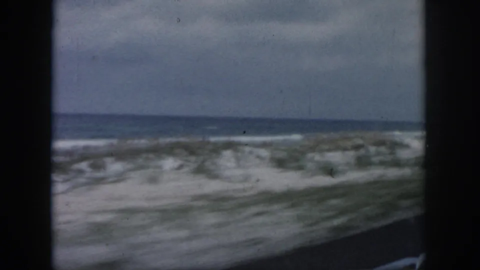 1958 8mm coastal travel frame from the New Orleans, Louisiana metadata set showing sandy beach, low dune vegetation, ocean water, and an overcast sky.