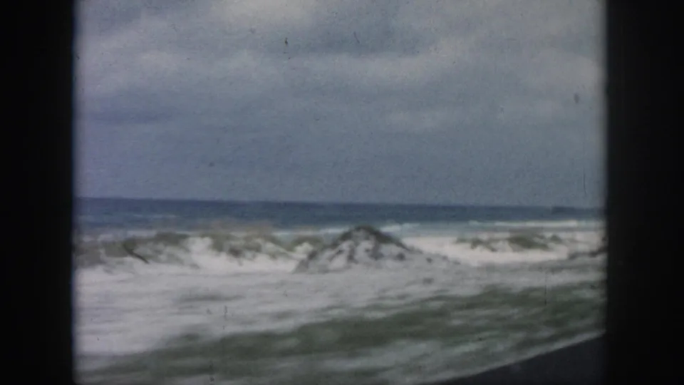 Vehicle-window view of breaking waves, pale sand, and low beach vegetation in the documented 1958 New Orleans coastal clip.