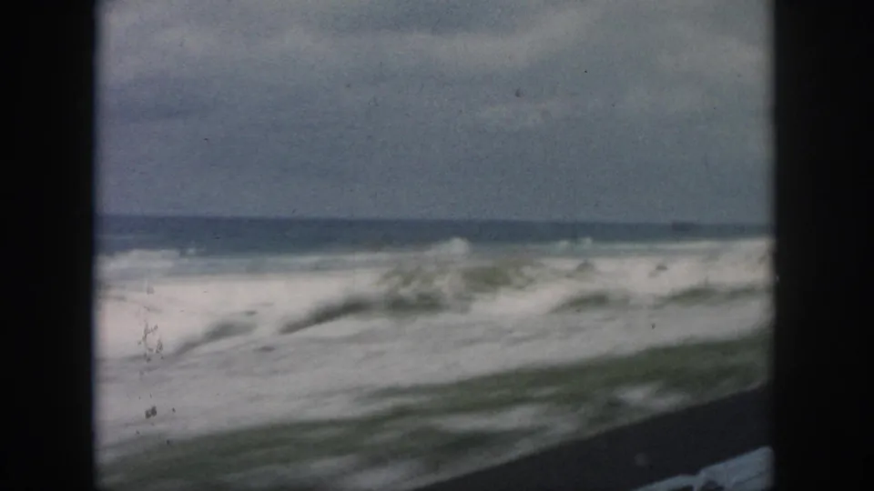 Soft 8mm frame of stormy-looking surf and sandy shoreline passing by from inside a moving vehicle in 1958 Louisiana footage.