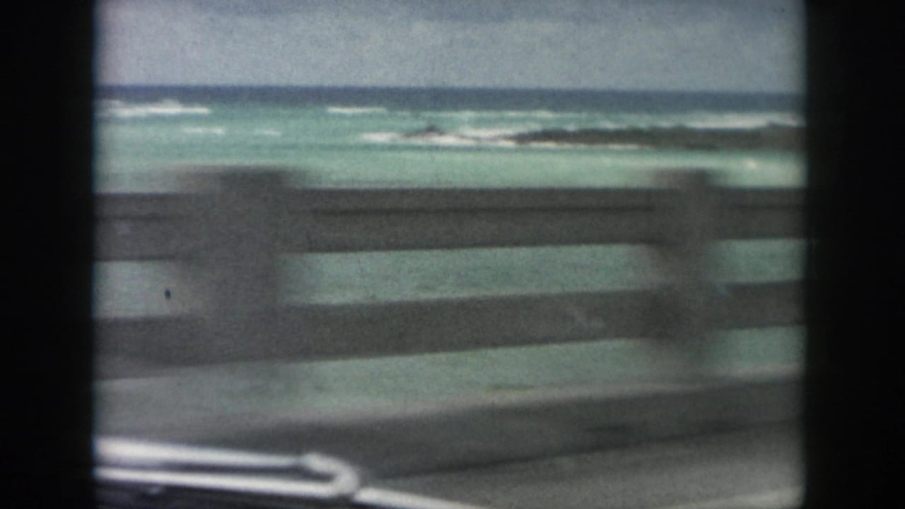 Roadside guardrail blurs past turquoise coastal water and low surf in 1958 Louisiana home-movie footage.