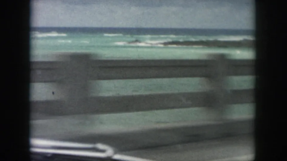 Roadside guardrail blurs past turquoise coastal water and low surf in 1958 Louisiana home-movie footage.