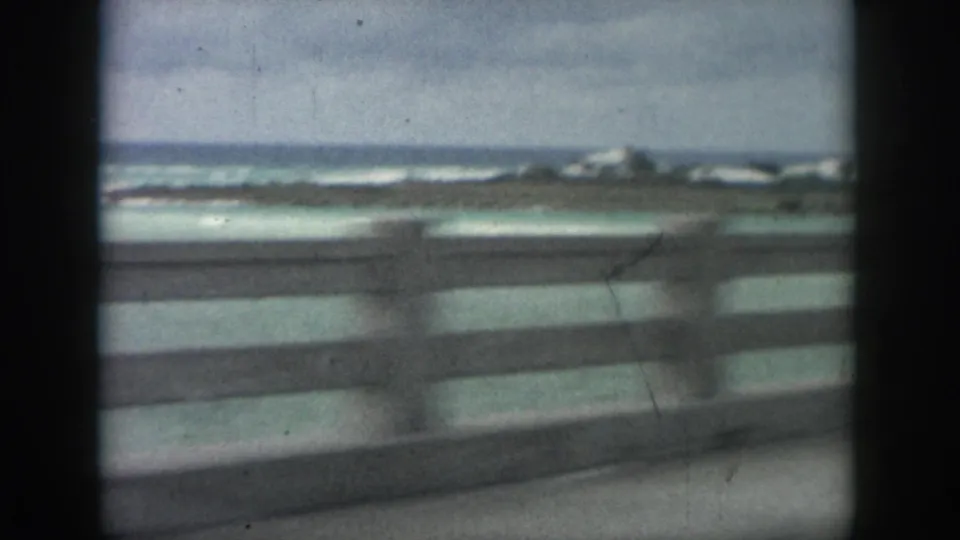 A moving-car view shows a coastal railing, turquoise water, and white surf softened by 8mm blur and faded color.