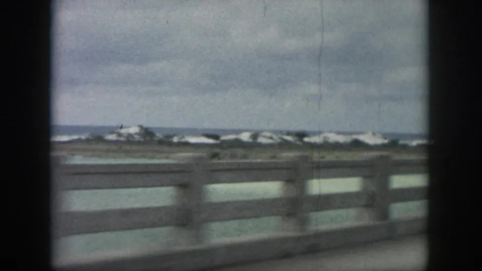 1958 home-movie frame of a coastal road guardrail, distant white-capped waves, and muted sky near New Orleans, Louisiana.