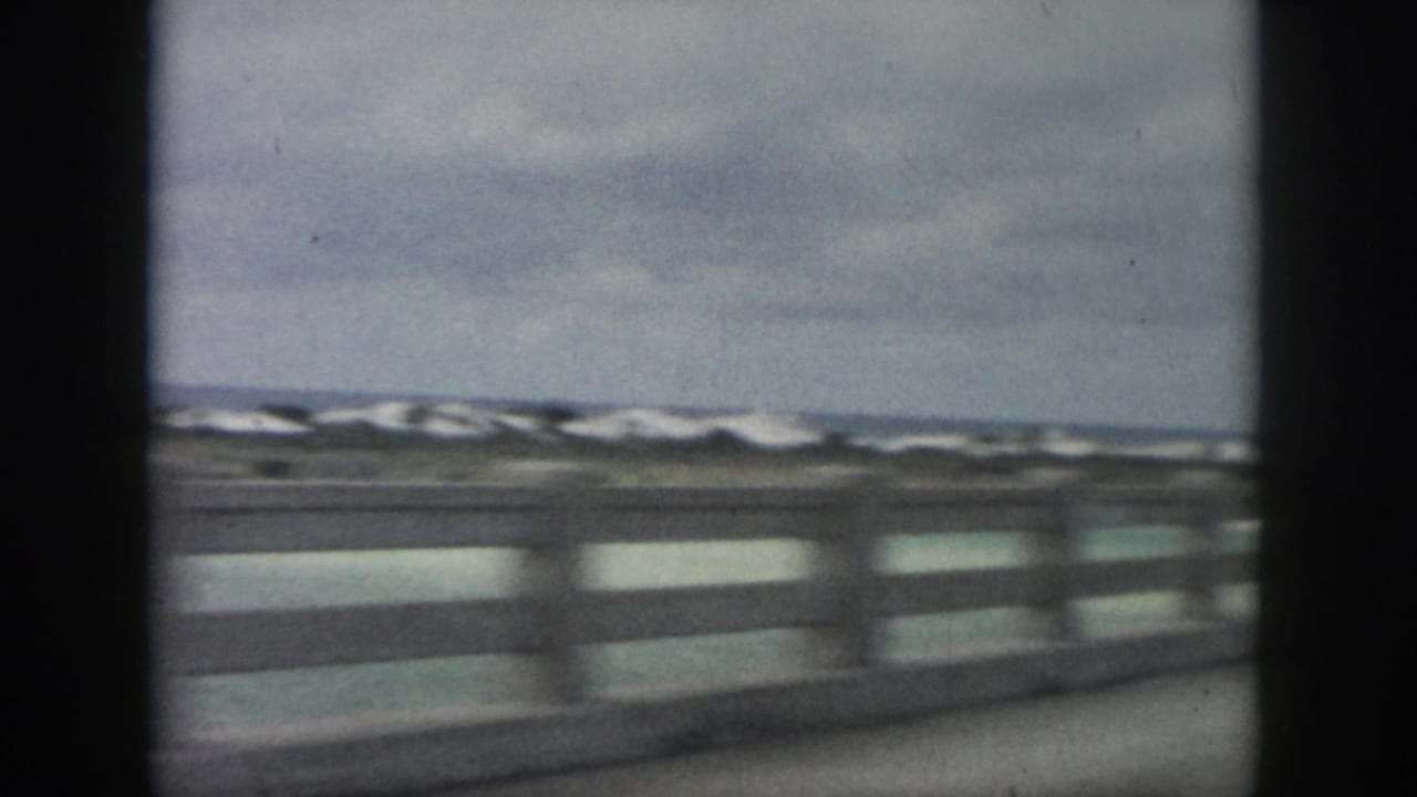 1958 New Orleans-area 8mm frame of a coastal road view, with concrete railing, pale dunes, and rough water beyond.