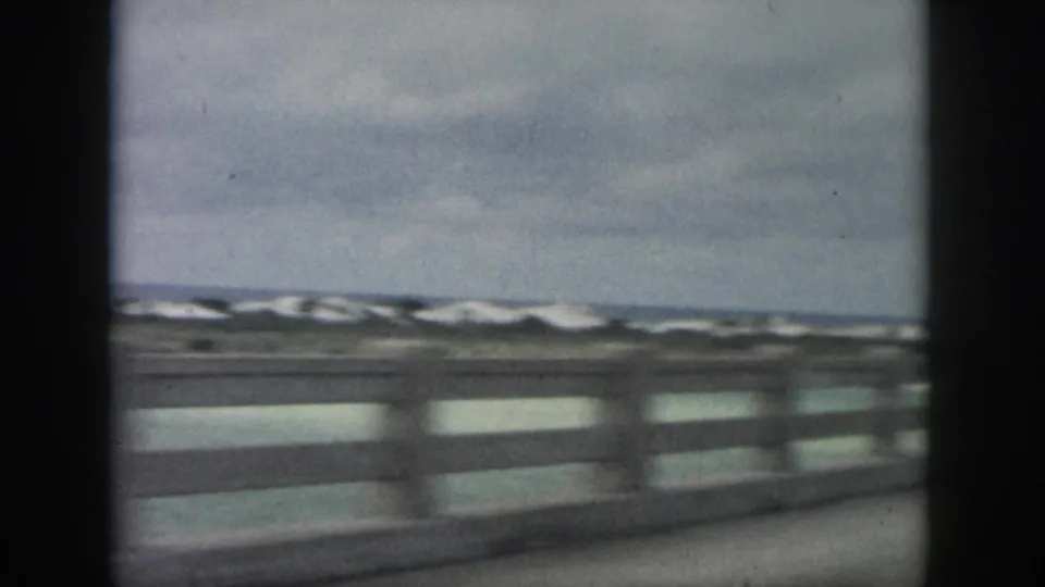 1958 New Orleans-area 8mm frame of a coastal road view, with concrete railing, pale dunes, and rough water beyond.