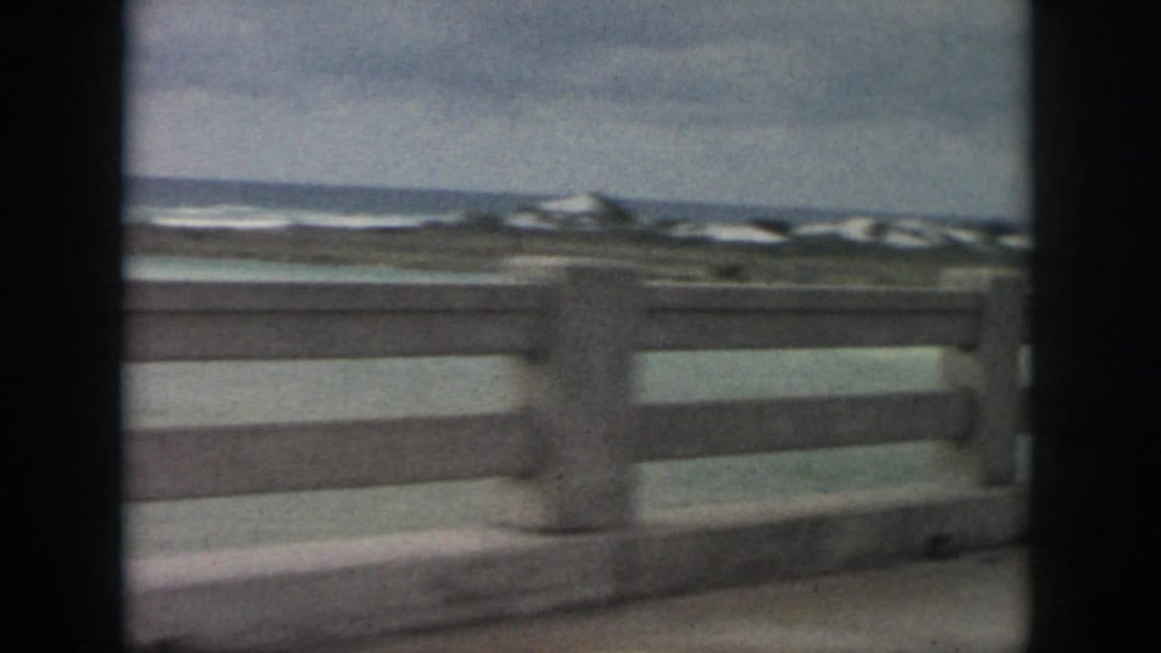 A moving 8mm view along a New Orleans-area coastal road, showing railing, sea, low shore, and cloudy sky.