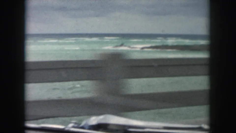 1958 8mm coastal drive frame with turquoise surf, a concrete roadside barrier, and a glimpse of the car hood.
