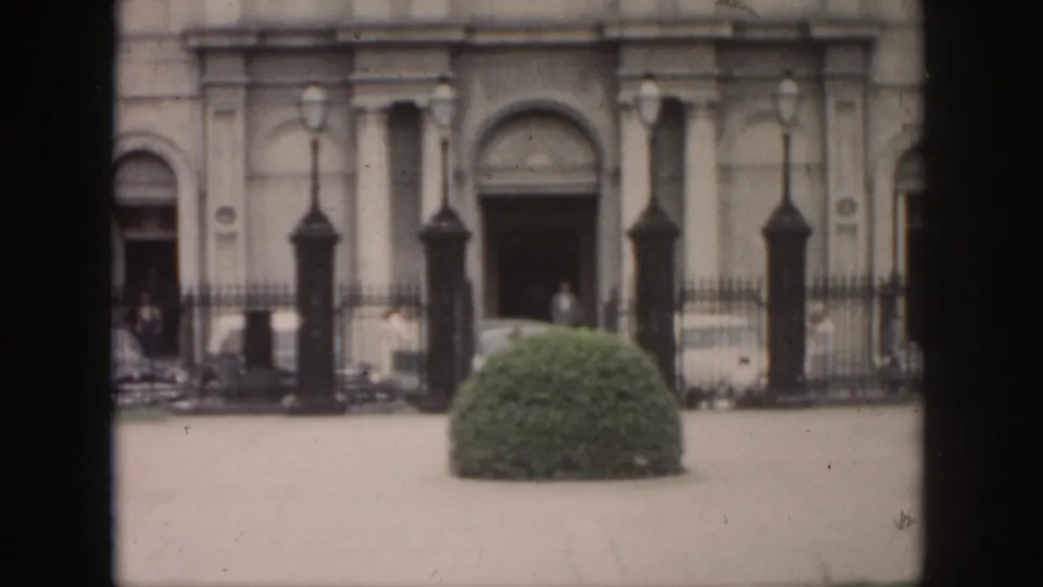 New Orleans 1958 home-movie frame showing an arched building frontage with lampposts, a fence, shrub, and distant people.