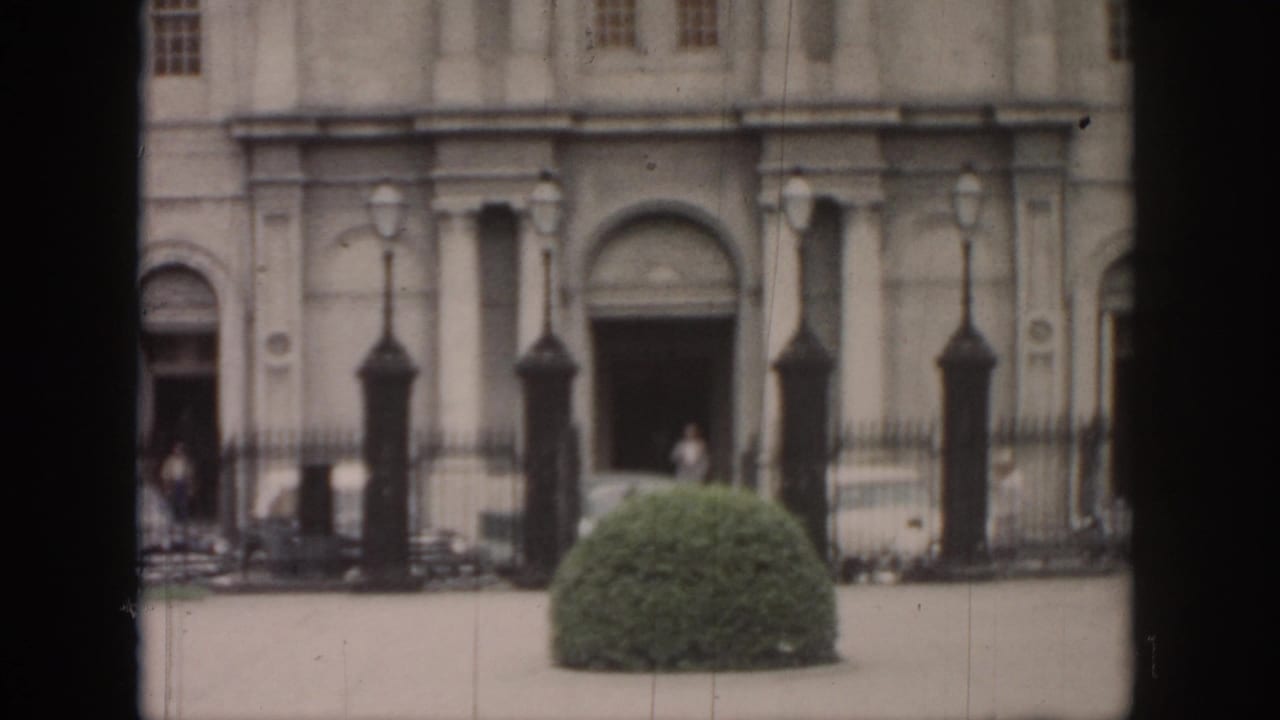 Pale classical building facade with iron fence, lamps, shrub, and small figures near the entrance.