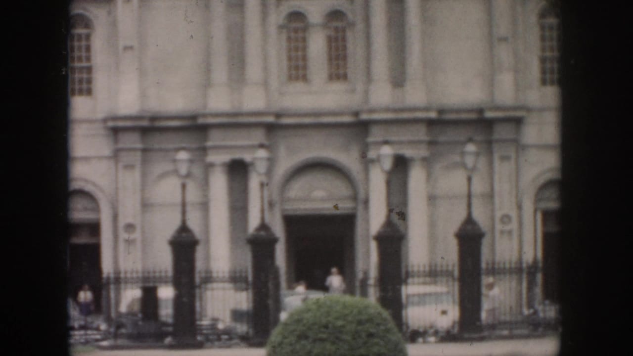 Classical building entrance with columns, arched doorway, fence, lamps, shrub, and pedestrians.