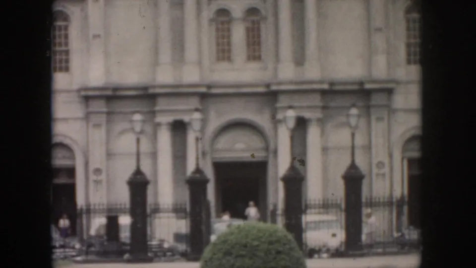 1958 New Orleans exterior view of a formal building entrance with columns, arched doorway, iron fence, lamps, and people nearby.