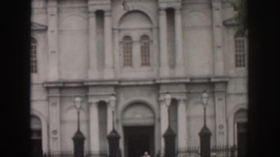 1958 New Orleans 8mm frame looking up the tall classical facade, with upper windows, entrance, lamps, fence, and distant pedestrians.