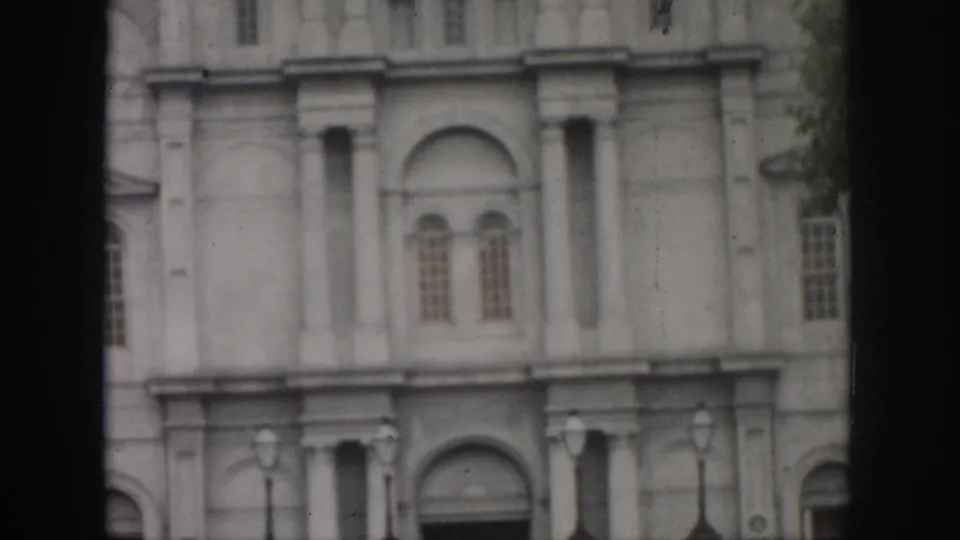 1958 New Orleans home-movie frame of the tall pale building facade, showing arched doorway, windows, iron fence, lamps, and a tree edge.