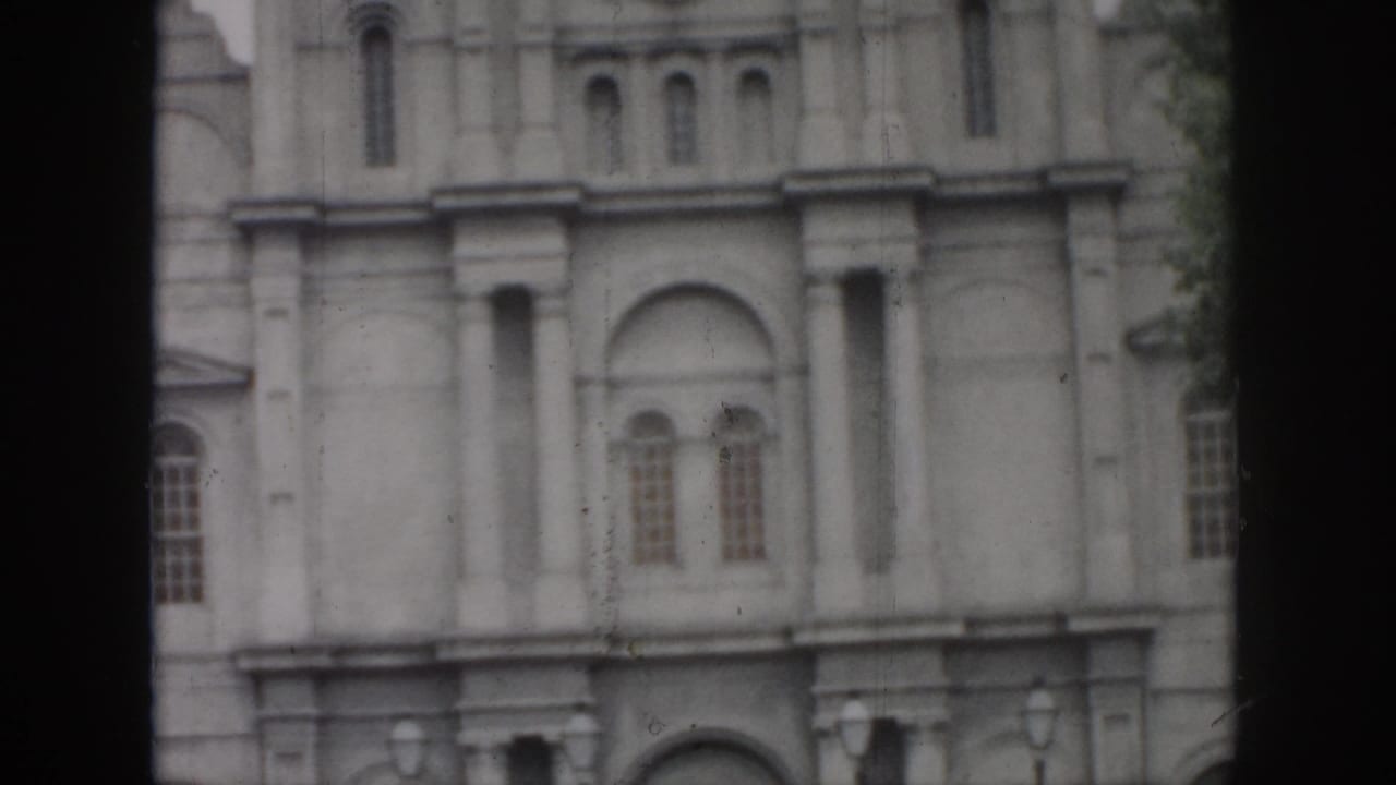 1958 New Orleans home-movie frame showing a pale masonry facade with arched windows, columns, and dark film borders.