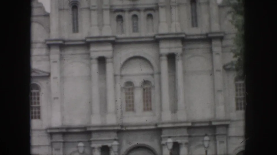 1958 New Orleans home-movie frame showing a pale masonry facade with arched windows, columns, and dark film borders.