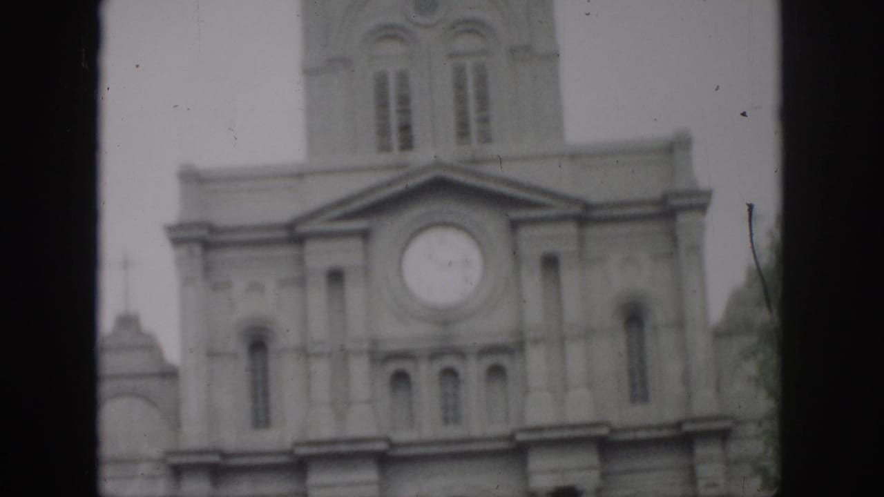 1958 New Orleans home-movie frame showing a church facade with a round clock, arched windows, and tower through soft 8mm focus.