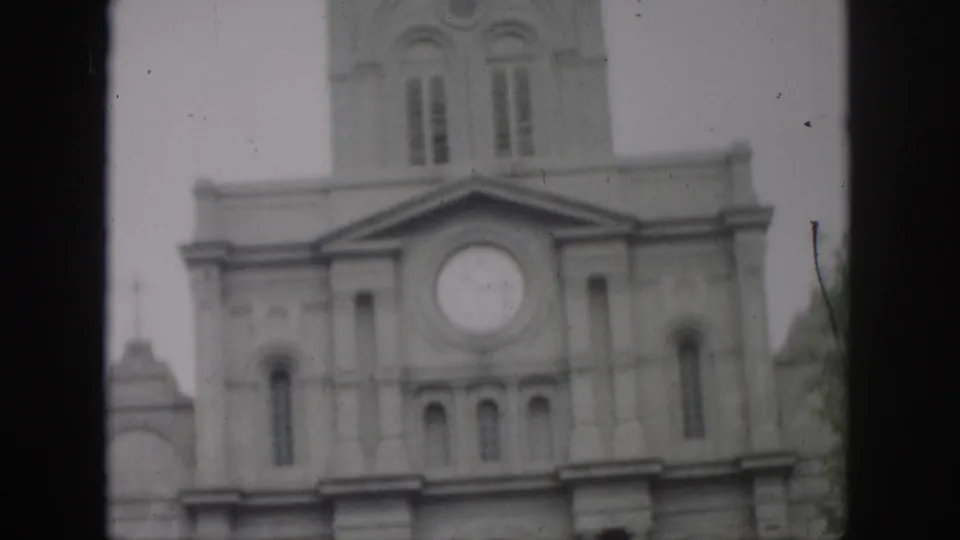 1958 New Orleans home-movie frame showing a church facade with a round clock, arched windows, and tower through soft 8mm focus.