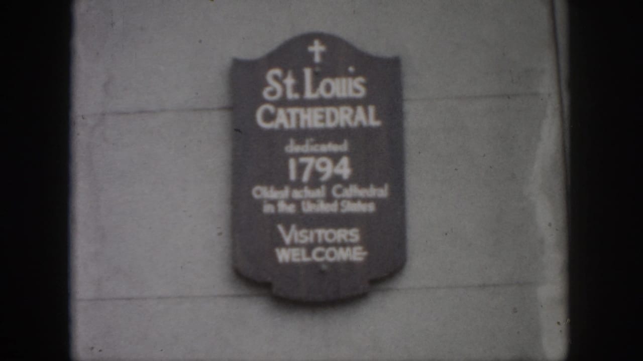 1958 New Orleans home-movie frame showing a St. Louis Cathedral plaque mounted on a stone wall, softly focused and grainy.