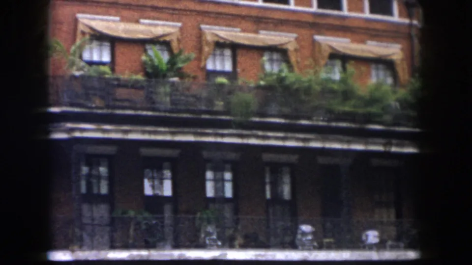 Grainy 1958 New Orleans frame showing a brick facade with balcony plants, iron railings, awnings, and shadowed lower openings.