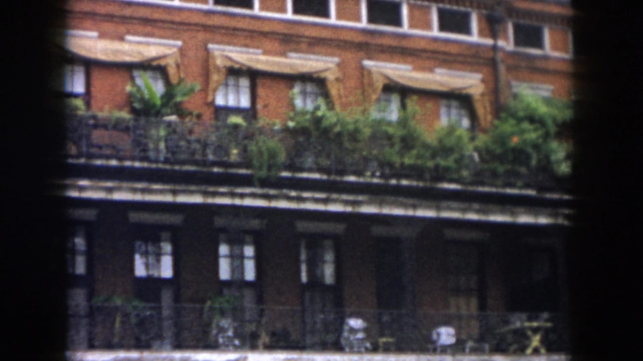 1958 New Orleans home-movie frame showing a red brick facade with windows, awnings, balcony plants, and dark vignetted edges.