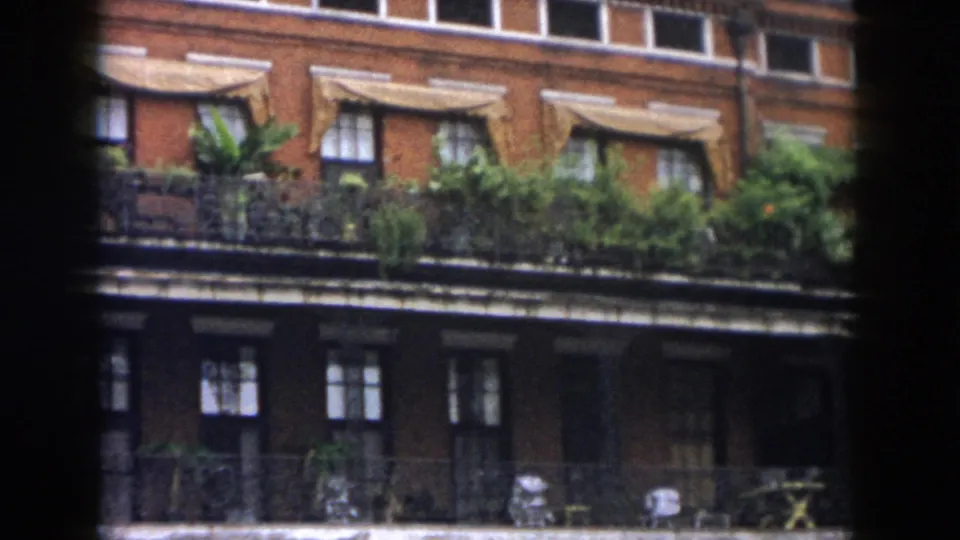 1958 New Orleans home-movie frame showing a red brick facade with windows, awnings, balcony plants, and dark vignetted edges.
