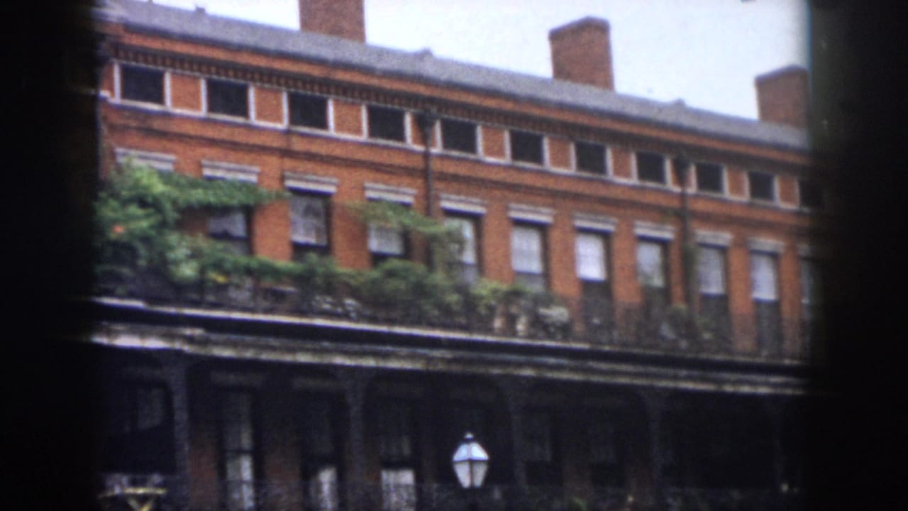 1958 New Orleans home-movie frame showing a red brick facade with tall windows, wrought-iron balconies, ivy-like greenery, and a street lamp below.