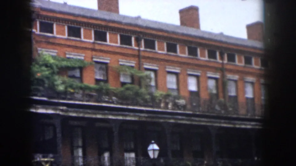 1958 New Orleans home-movie frame showing a red brick facade with tall windows, wrought-iron balconies, ivy-like greenery, and a street lamp below.