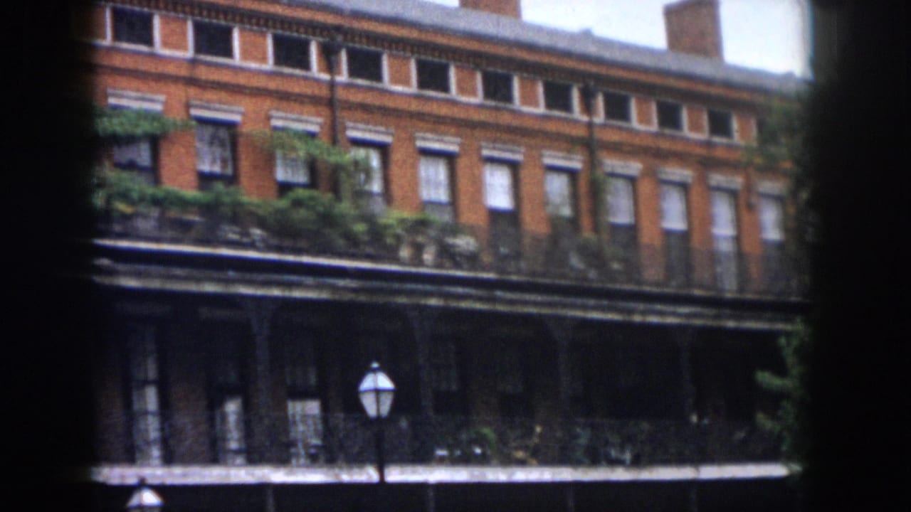 Brick facade with upper windows, leafy balcony plants, dark porch level, and a lamp post.