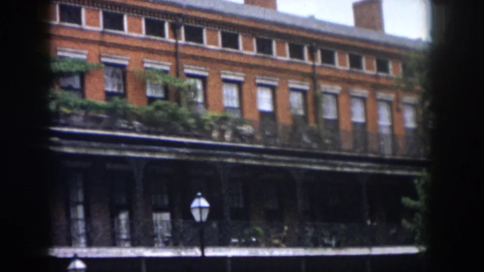 1958 New Orleans exterior frame with red brick rows, white window trim, planted balcony railings, a shaded lower gallery, and a traditional lamp.