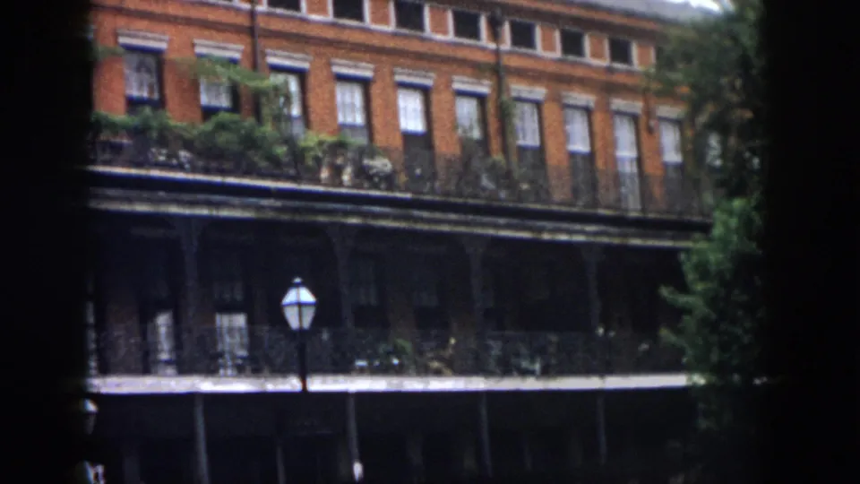 1958 New Orleans home-movie view of a brick building facade with iron balcony railings, greenery, a courtyard lamp, and tree foliage along the edge.