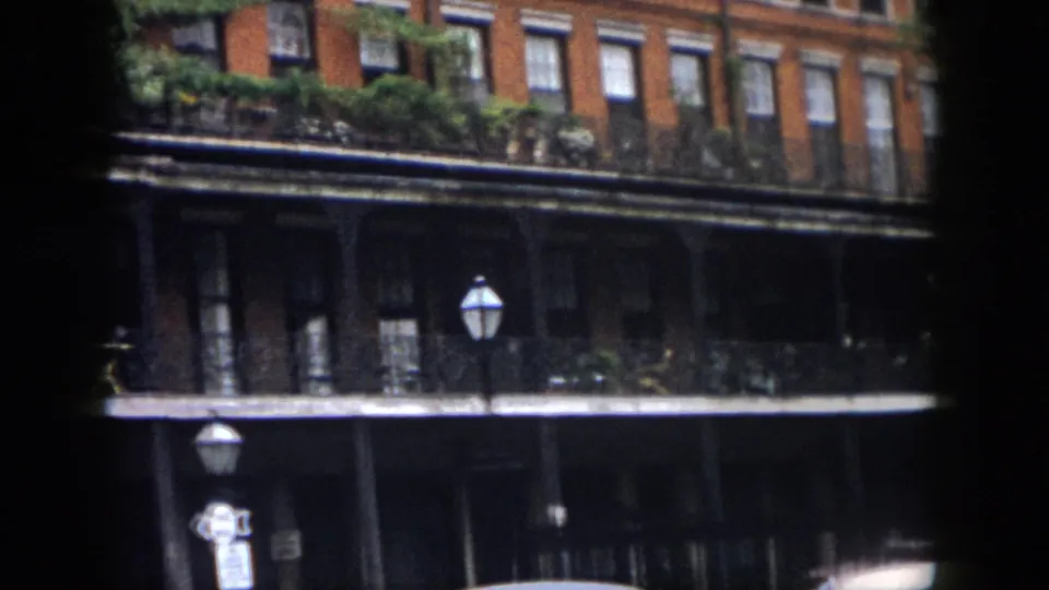 1958 New Orleans exterior frame showing a red brick facade, wrought-iron galleries, balcony greenery, street lamps, and dark lower-level openings.