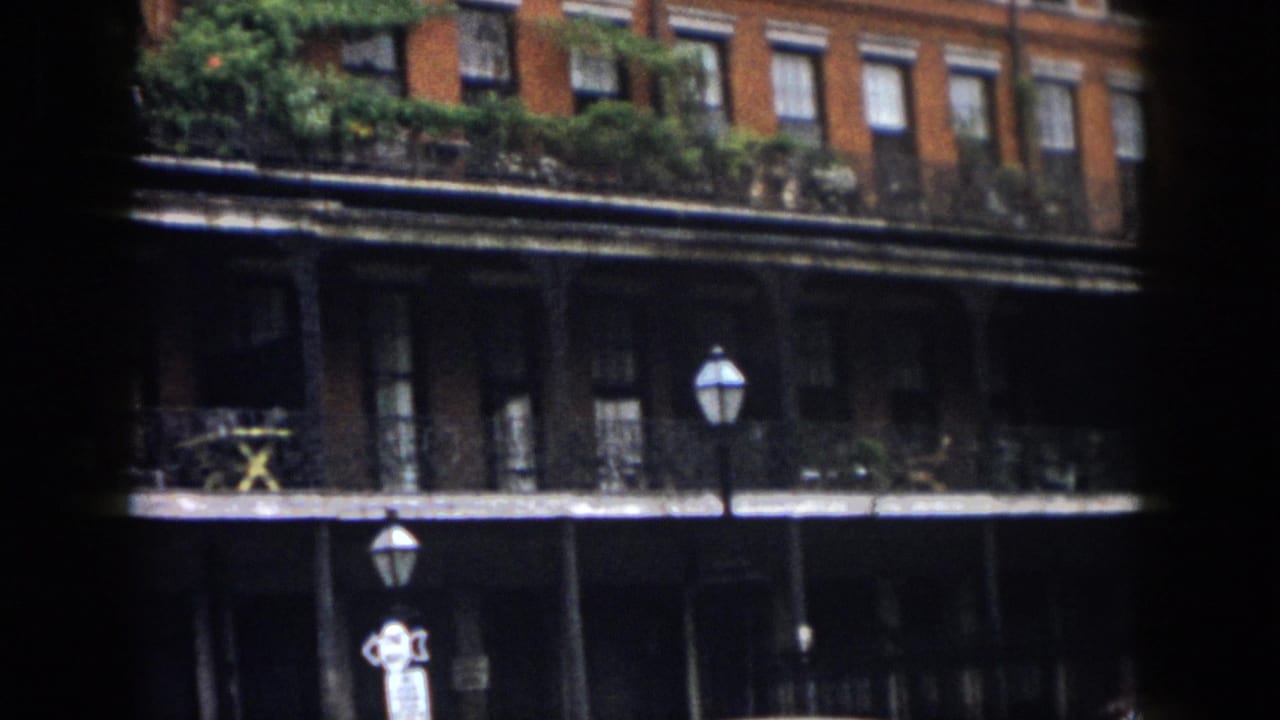 1958 New Orleans 8mm frame showing a brick facade, wrought-iron balcony, plants, and street lamps seen through dark edges.