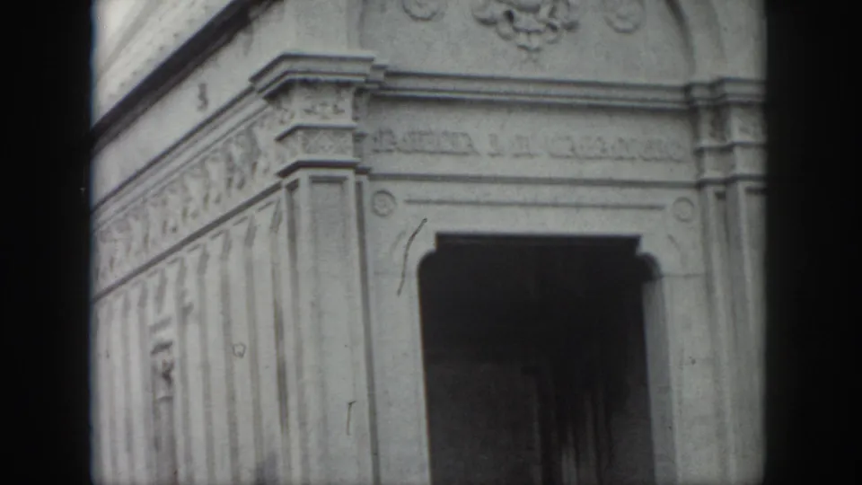 Mid-century 8mm frame from New Orleans showing carved stone panels, a mausoleum doorway, and the softened contrast of black-and-white film.