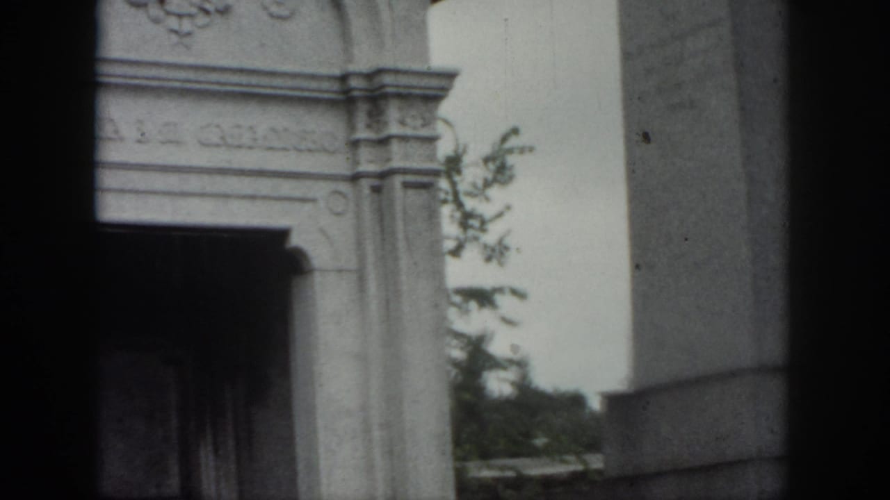 New Orleans cemetery frame showing part of the mausoleum entrance, a large stone monument, and greenery behind it.
