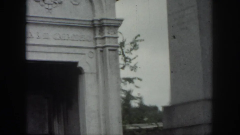 New Orleans cemetery frame showing part of the mausoleum entrance, a large stone monument, and greenery behind it.