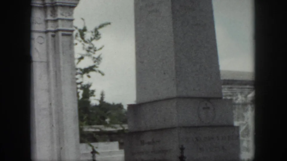 1958 cemetery shot with the base of a stone obelisk, the edge of a mausoleum, distant tombs, and trees.