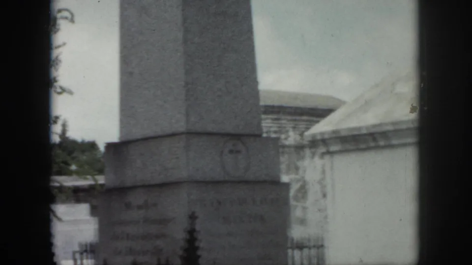 New Orleans cemetery view of an obelisk base marked with a cross emblem, with iron fencing and another tomb nearby.