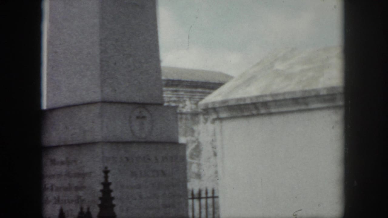 1958 New Orleans cemetery view with a tall stone monument, faint inscriptions, iron fencing, and pale mausoleum roofs in soft 8mm focus.