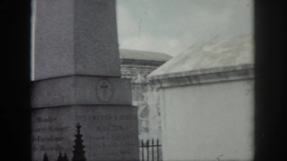 1958 New Orleans cemetery view with a tall stone monument, faint inscriptions, iron fencing, and pale mausoleum roofs in soft 8mm focus.