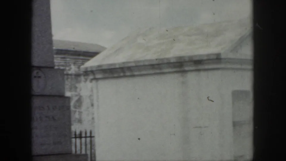 1958 New Orleans cemetery frame showing a vertical stone monument with a cross emblem, pale mausoleum rooflines, and iron fencing.