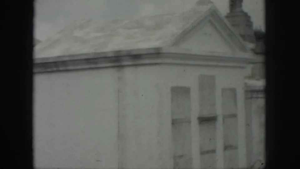 1958 New Orleans cemetery view of a pale mausoleum with a triangular pediment, chimney-like detail, and soft black-and-white 8mm texture.