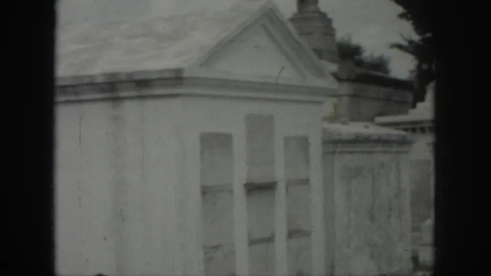 1958 New Orleans cemetery frame showing a pale mausoleum facade, recessed window shapes, a pedimented roofline, and nearby stonework.