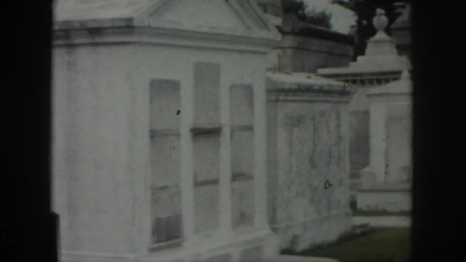 1958 New Orleans home-movie view of pale cemetery mausoleums and stone walls with soft focus and dark film edges.