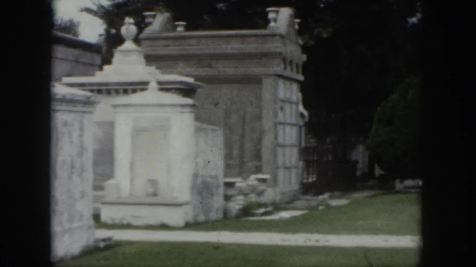 1958 New Orleans visitor footage of a cemetery path with white stone monuments, lawn, and a manicured tree.