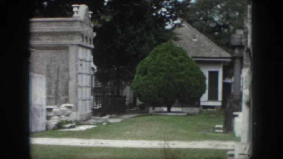 1958 New Orleans cemetery view with above-ground stonework, clipped greenery, path, and a small white building beyond.